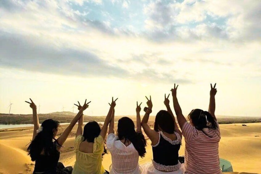 A group of people sitting on a sandy beach with their arms raised, facing a cloudy sky. 1