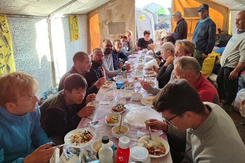A group of people seated at a long table, eating and conversing in a tented area. 2