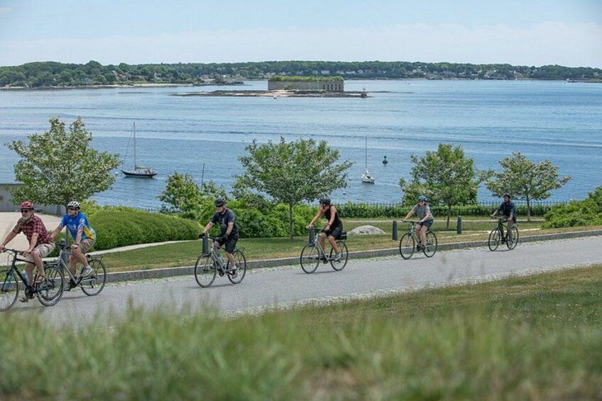 A group of cyclists on a paved path by a body of water with trees and a distant shoreline. 1