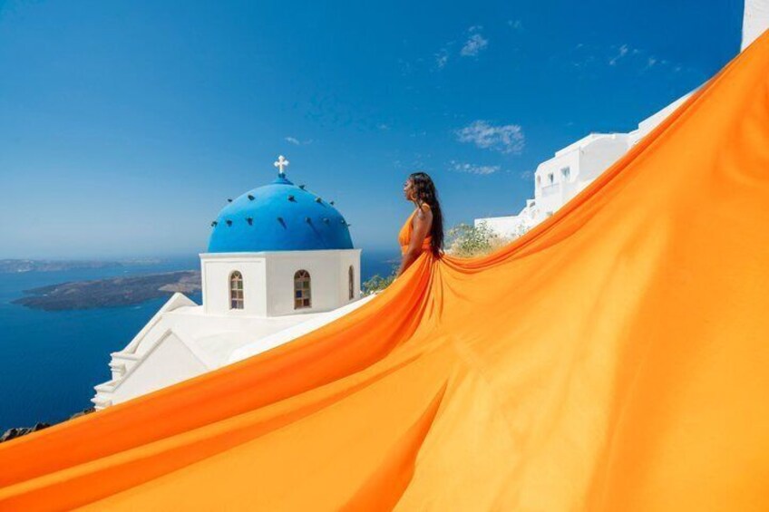 A woman in an orange dress standing on a cliff with a white building and blue dome in the background. 5