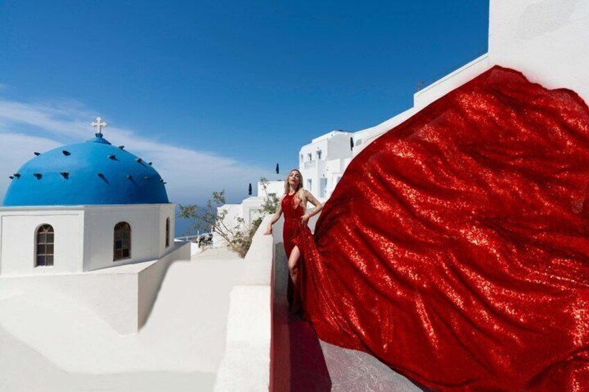 A person in a red dress leaning on a white wall with a blue dome and white buildings in the background. 1