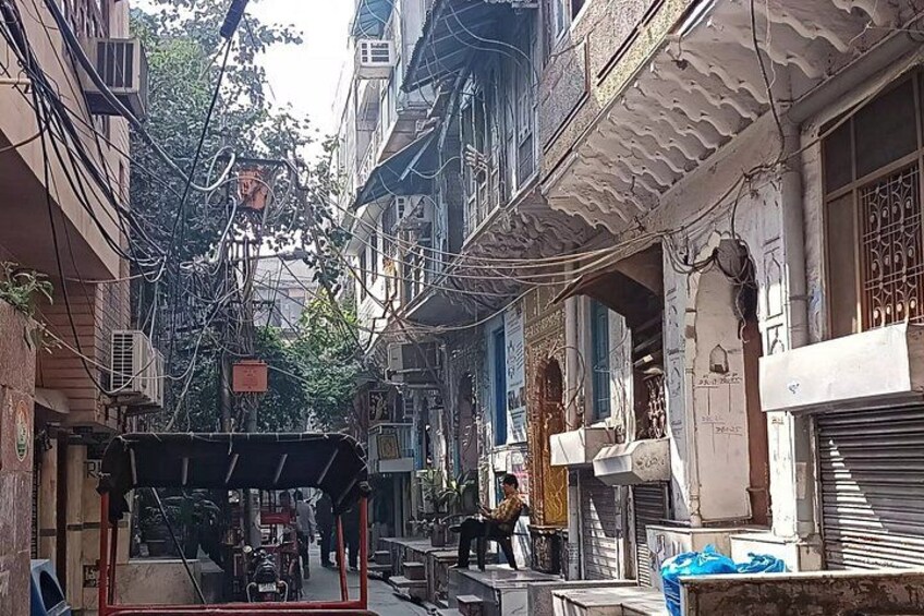 A narrow street with buildings on either side, some with balconies and air conditioning units. There are visible electrical wires and a small structure in the foreground. 5