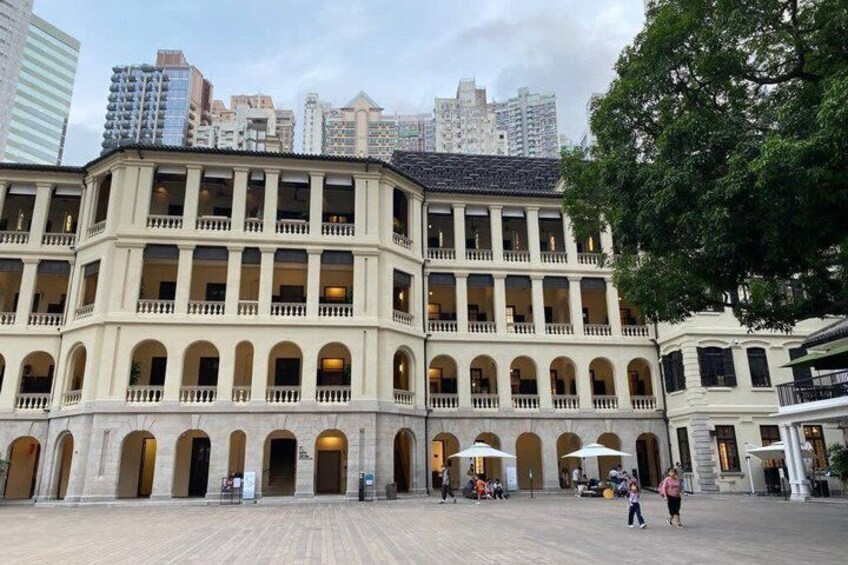 A historic building with multiple arches and balconies, surrounded by a plaza with people walking. 1