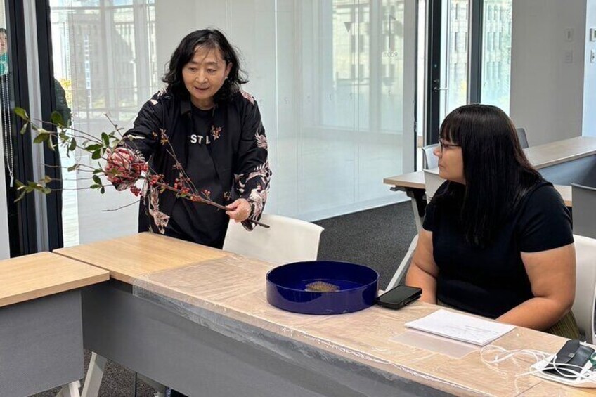 Two people at a desk with a blue container, a plant, and a laptop. 4