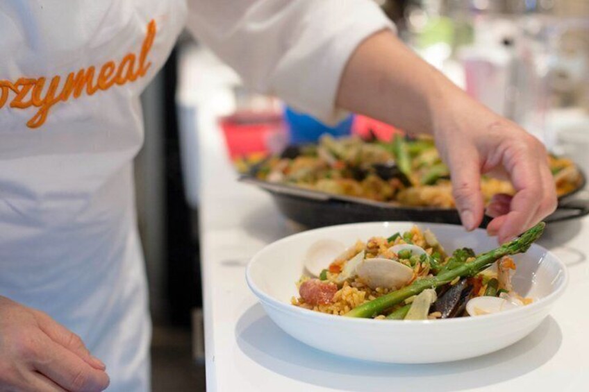 A person in a white apron with 'OZYMEL' text serving food from a pan to a plate. 1