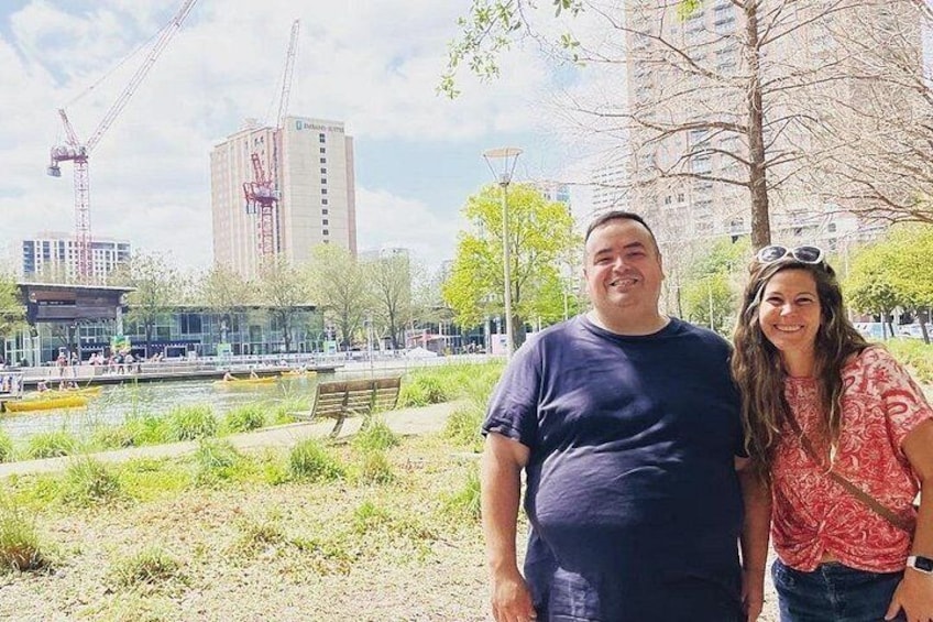 A man and a woman standing in a park with a river, trees, and buildings in the background. 5