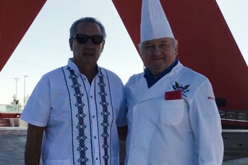 Two individuals in white chef uniforms with black embroidery, standing in front of a red structure. 5