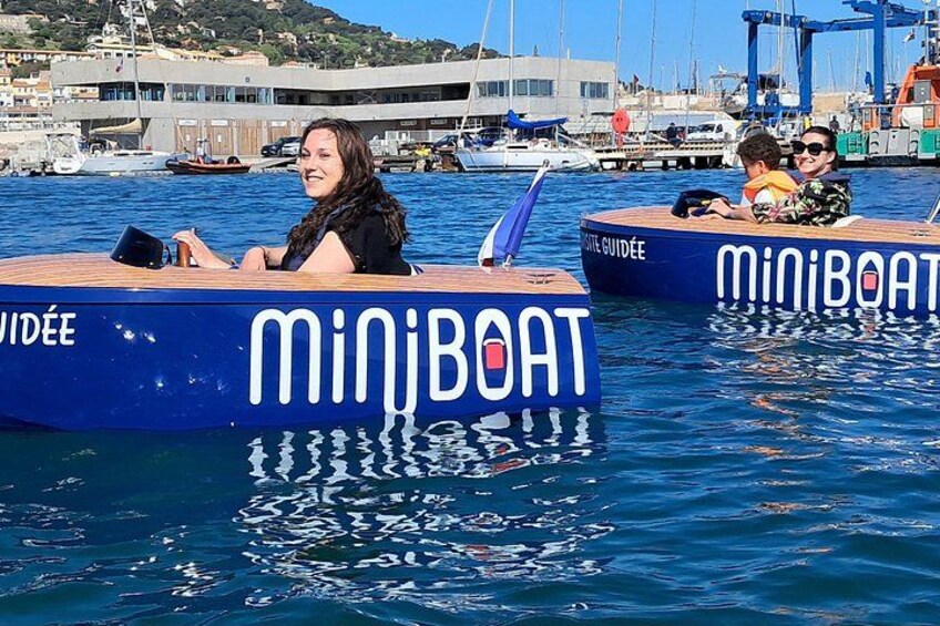 Two blue boats with 'MiniBoat' written on them, docked at a marina with buildings and cranes in the background. 3