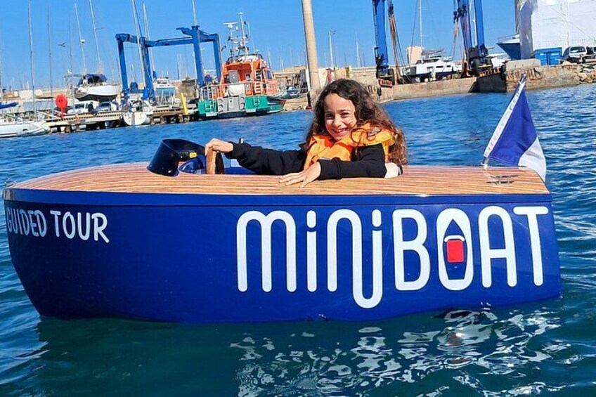 A person in a boat with 'MINIBOAT' written on it, docked at a marina with other boats and a flag. 4