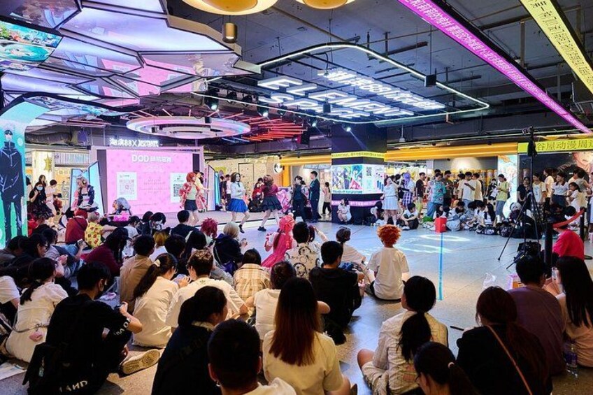 A crowded indoor event with a stage, audience, and various booths. 2