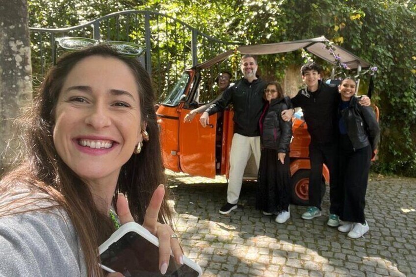 A group of people posing in front of a vintage orange car with a canopy, surrounded by greenery. 4