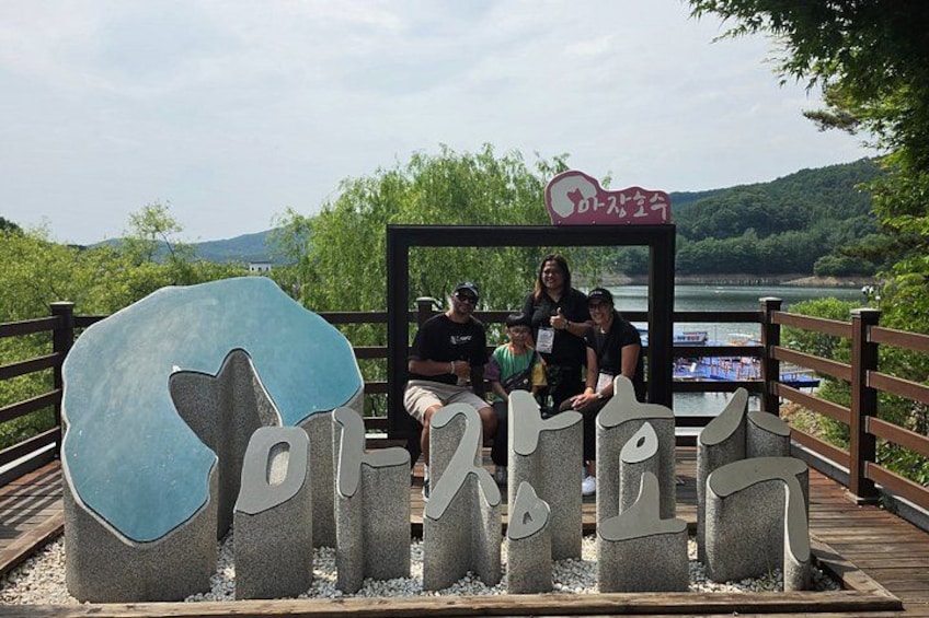 Eine Gruppe von Menschen sitzt auf einer Holzplattform, im Vordergrund ein großes blau-weißes Schild und im Hintergrund ein Fluss. 2