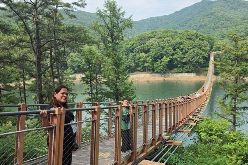 Un pont suspendu en bois enjambant une rivière, entouré d’une forêt dense et de collines. 3