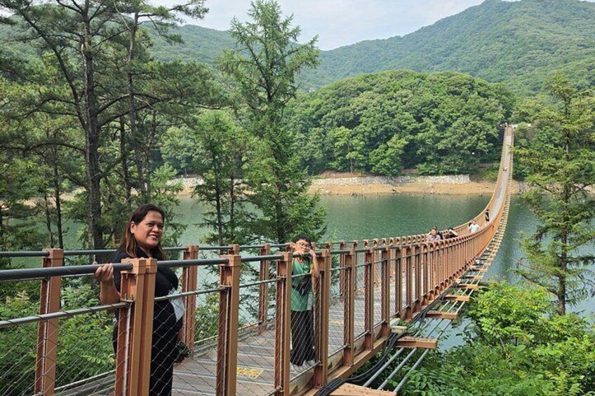 Eine hölzerne Hängebrücke über einem Fluss, umgeben von dichtem Wald und Hügeln. 2
