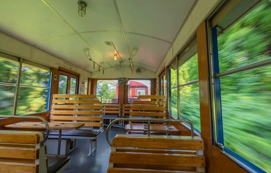 A train interior with wooden benches and metal tables, large windows, and a red train visible outside. 4