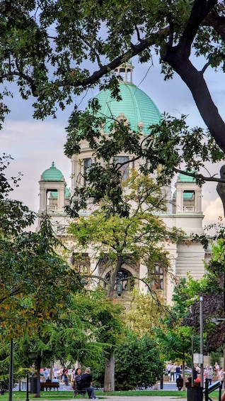 A historic building with a green dome and two smaller domes, surrounded by trees and a park with people. 3
