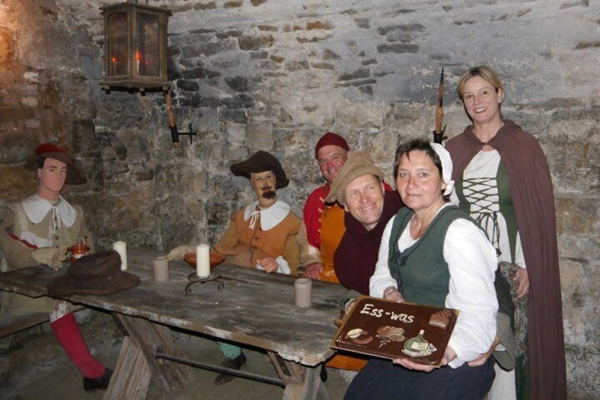 A group of people in historical costumes seated around a wooden table with candles, in a stone-walled room. 4