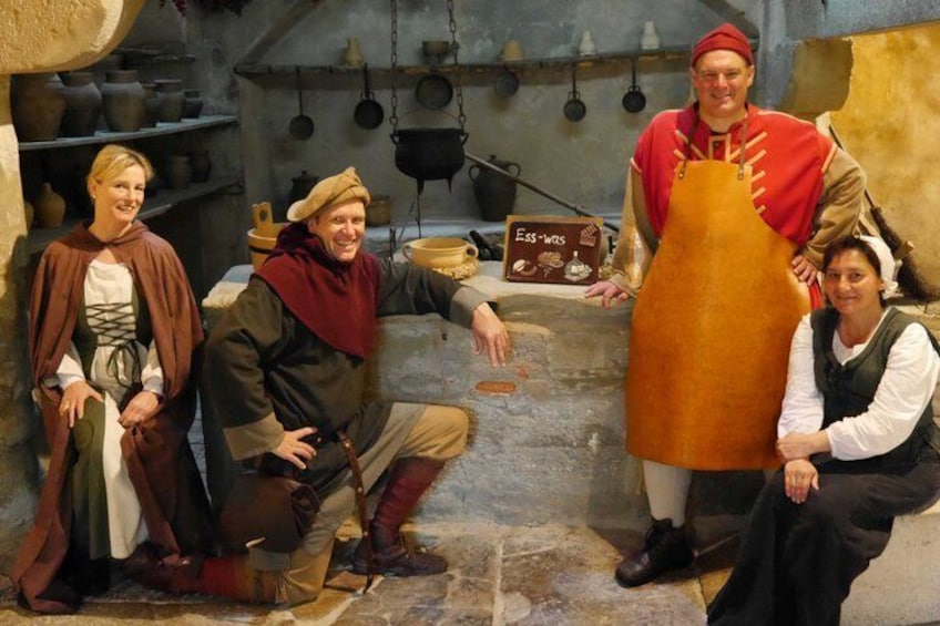 A historical reenactment scene with individuals in period costumes, a stone wall, and cooking utensils. 1