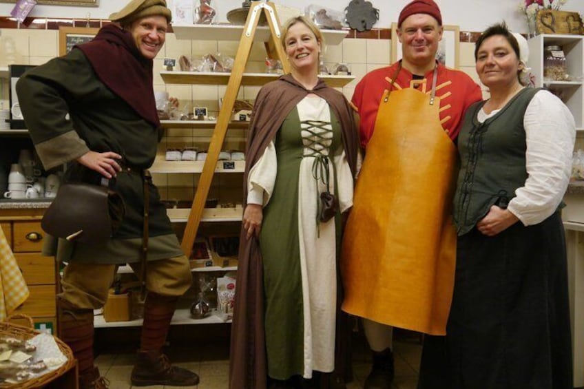 Four people in historical costumes in a kitchen setting with shelves and kitchenware. 3