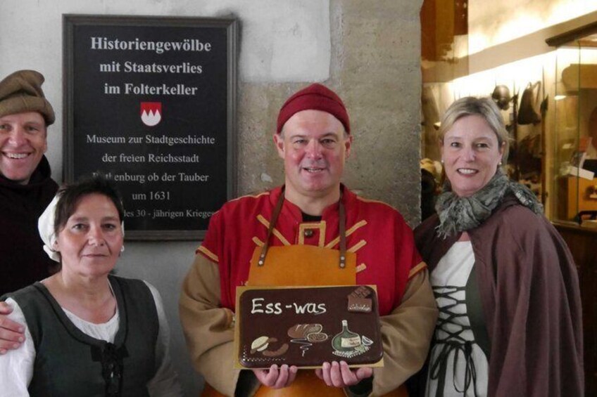 A historical exhibit with a sign, a person in traditional attire holding a book, and a woman in period clothing. 5