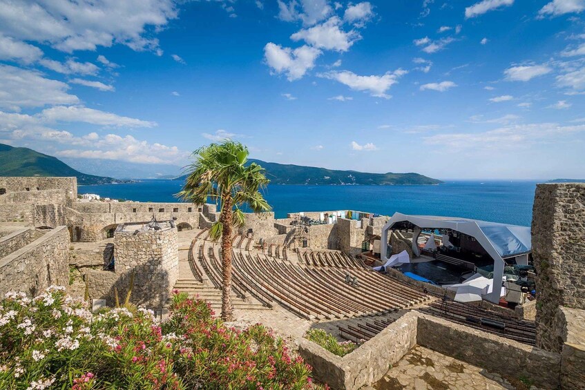 An ancient amphitheater with a modern stage, palm trees, and a coastal view. 4