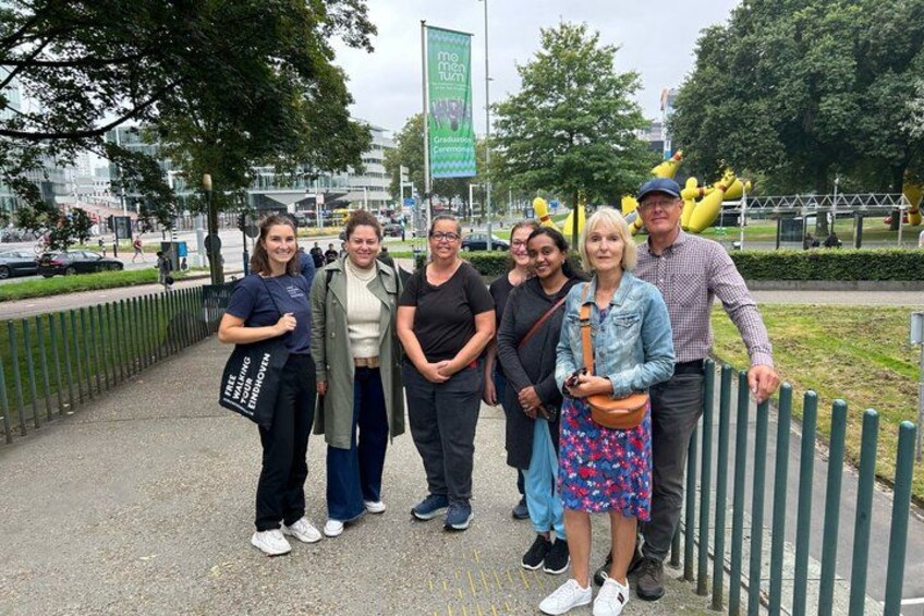 Un groupe de personnes se tient sur un trottoir, avec une clôture verte et des arbres en arrière-plan. 2