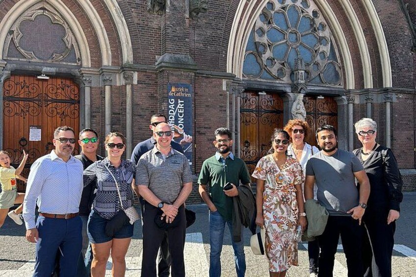 A group of people standing in front of a church with a large arched window and a sign that reads 'Muziek in de Kathedraal.' 2