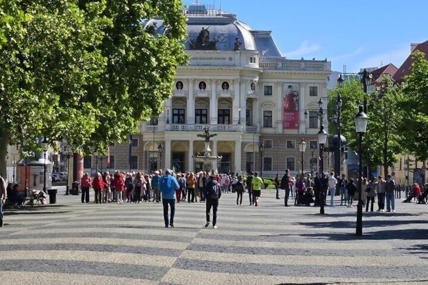 Una piazza pubblica con un grande edificio sullo sfondo, persone che passeggiano e alberi. 3
