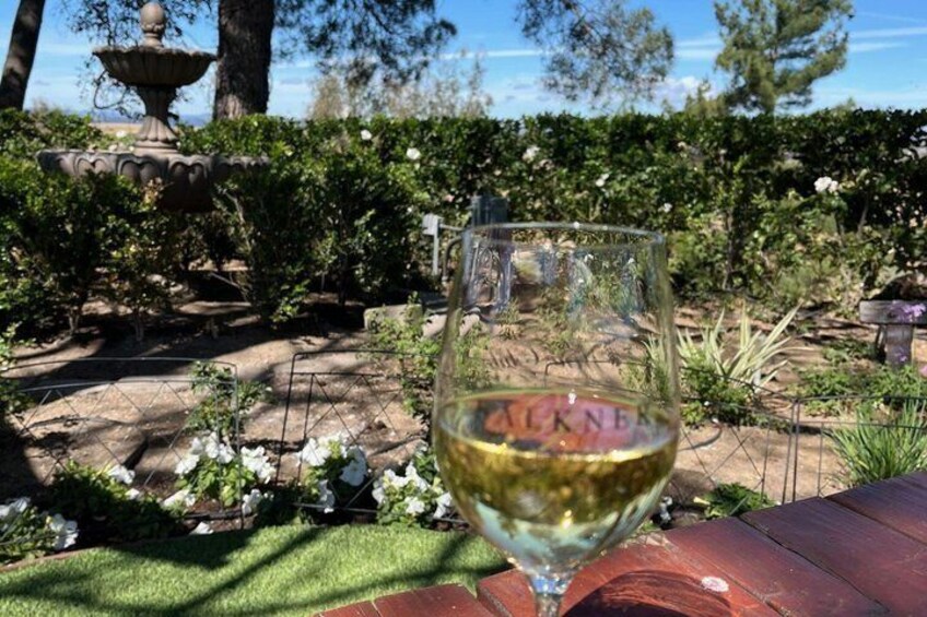A glass of white wine with 'ALKNE' on it, a garden with a fountain, and a wooden table. 5