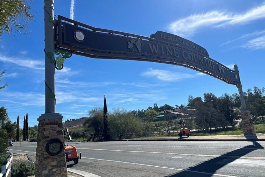 A road sign for Temecula Valley Wine Country with a clear blue sky and a red car on the road. 1