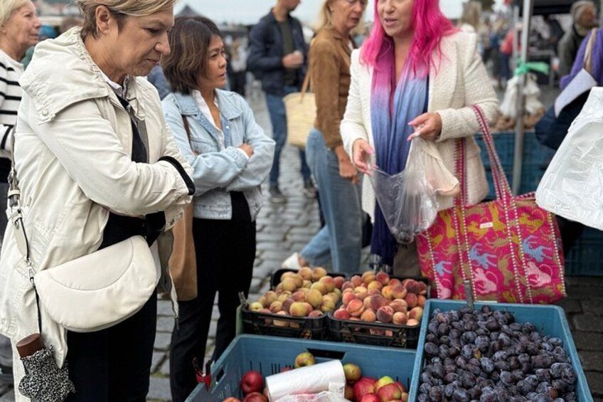 A market with people shopping, fruit stalls, and a cobblestone street. 2