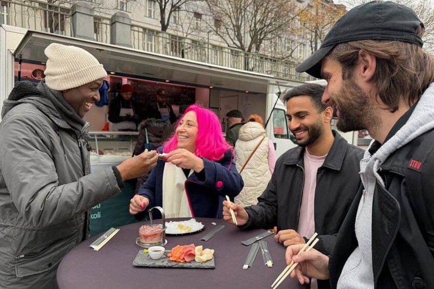 People dining at an outdoor food stall with a variety of dishes and utensils on the table. 2