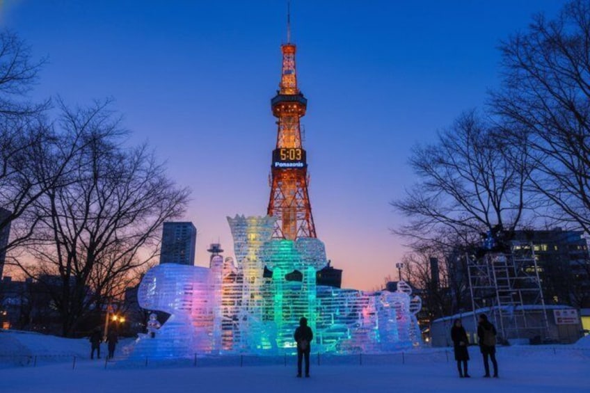 A brightly lit tower with a digital clock, surrounded by colorful ice sculptures and snow-covered ground. 3