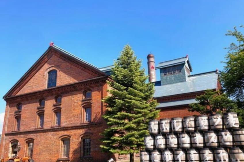 A red brick building with a large chimney, a tree in front, and a stack of barrels to the right. 2