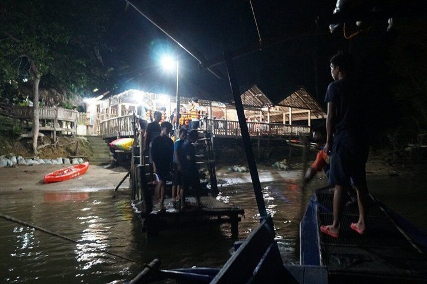 A nighttime scene with people on a boat, a red inflatable boat, and a building with a thatched roof in the background. 2