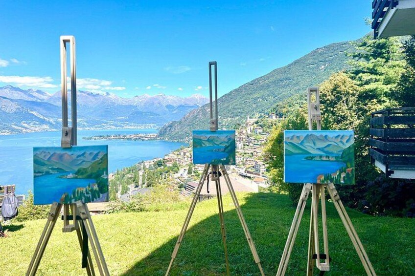 Three easels with landscape paintings, set against a backdrop of a lake, mountains, and a village. 1