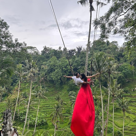 A person in a red dress swinging on a rope amidst lush green rice terraces and palm trees. 1