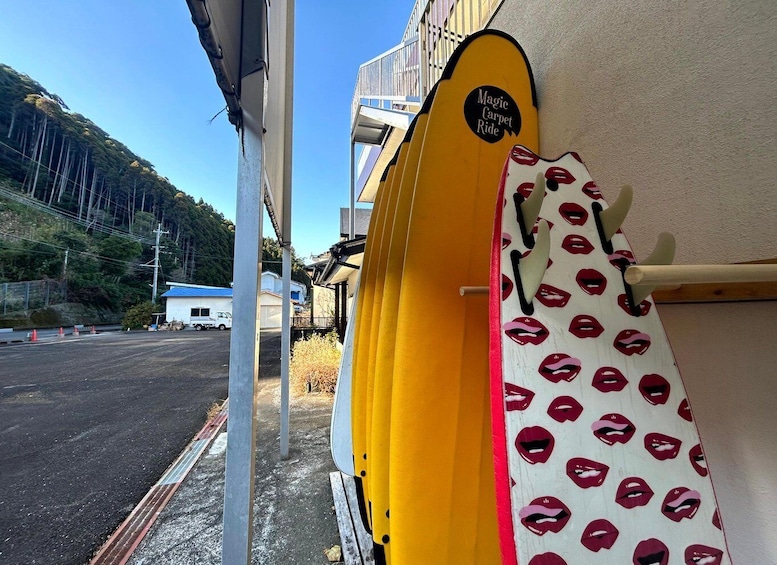 A yellow surfboard with 'Magic Carpet Ride' written on it, a white surfboard with red lips pattern, and a building with a blue roof in the background. 4