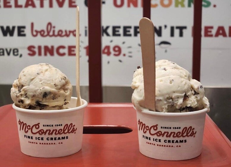 Two McConells Fine Ice Cream cups with wooden sticks, one with chocolate chips and one plain, in front of a sign with partially visible text. 5