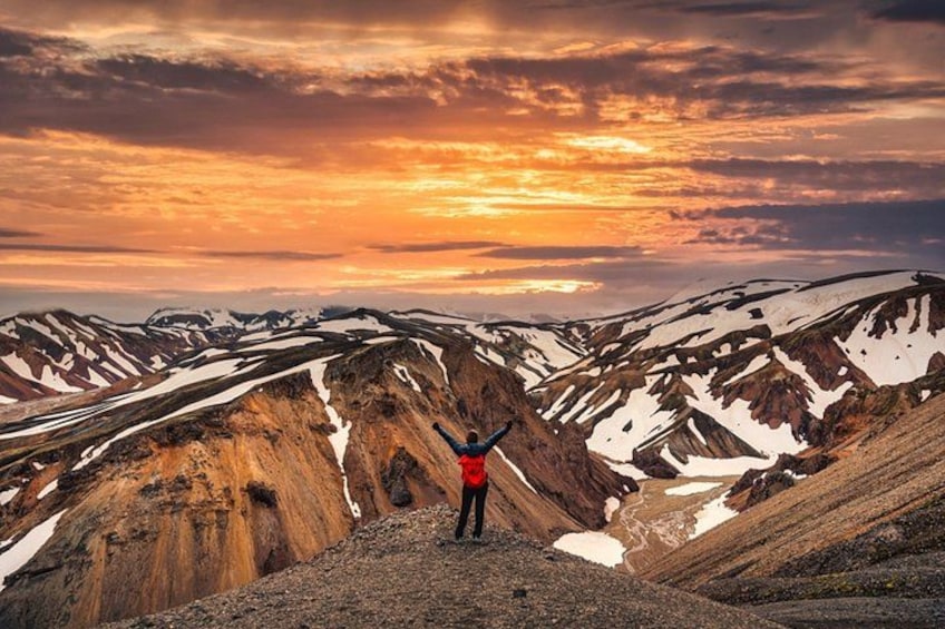 A person standing on a mountain ridge with arms raised, overlooking a valley with snow patches and a sunset sky. 1