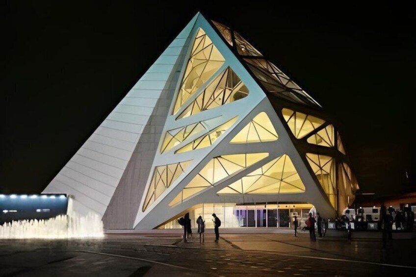 A modern, illuminated building with a triangular facade and a water feature in front. 3