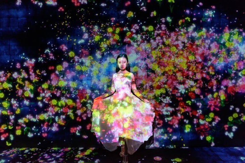 Eine Person in einem farbenfrohen Kleid mit Blumenmuster vor einem dunklen Himmel mit farbenprächtigen Blumenprojektionen. 1