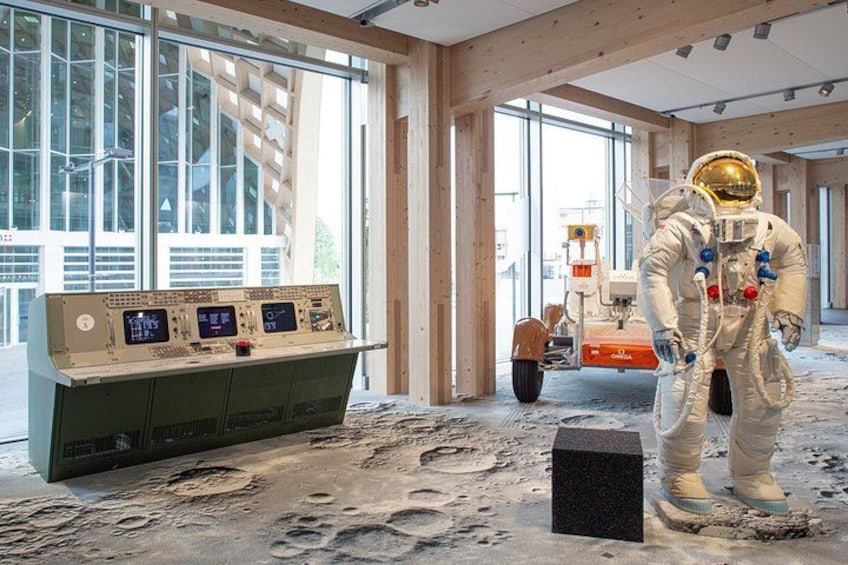 An astronaut in a space suit standing on a simulated lunar surface with a control panel and a construction vehicle in the background. 5