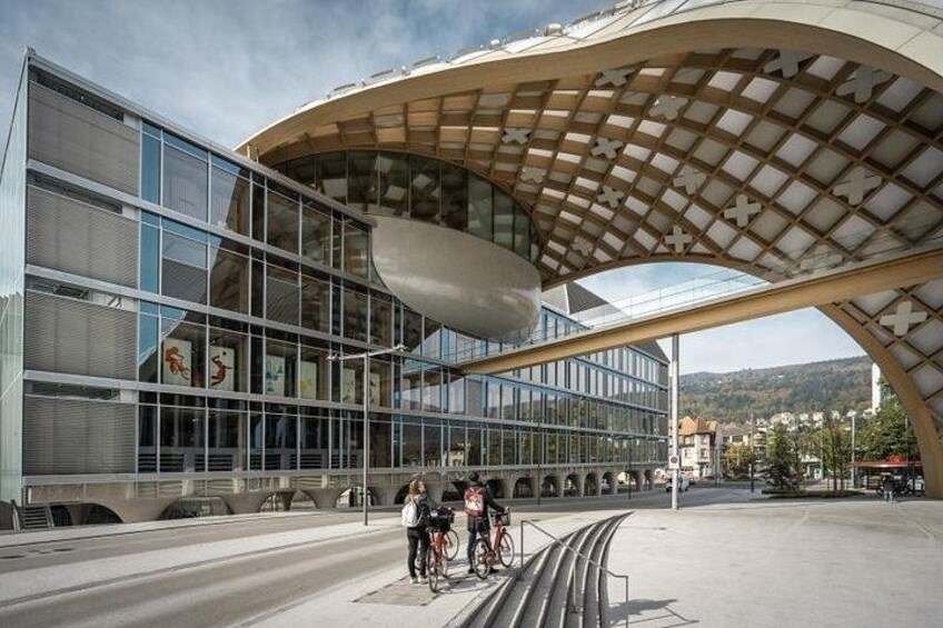A modern building with a curved glass facade and a wooden canopy structure. 1
