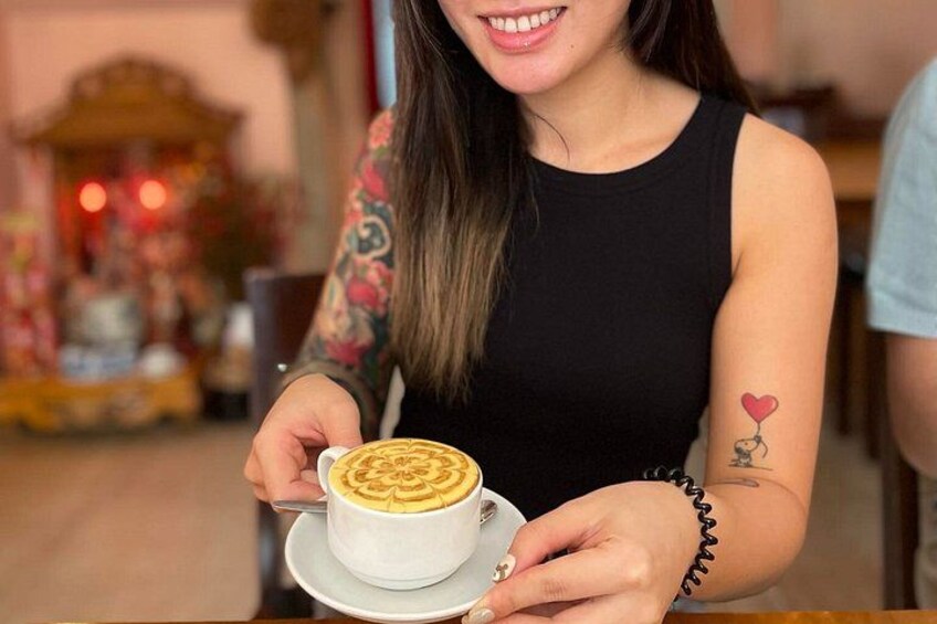 Une personne tenant une tasse de café décorée d’un motif en latte art, portant un débardeur noir et ayant un tatouage sur le bras. 3