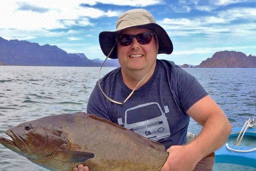 En person holder en stor fisk på en båd med bjerge i baggrunden. 2