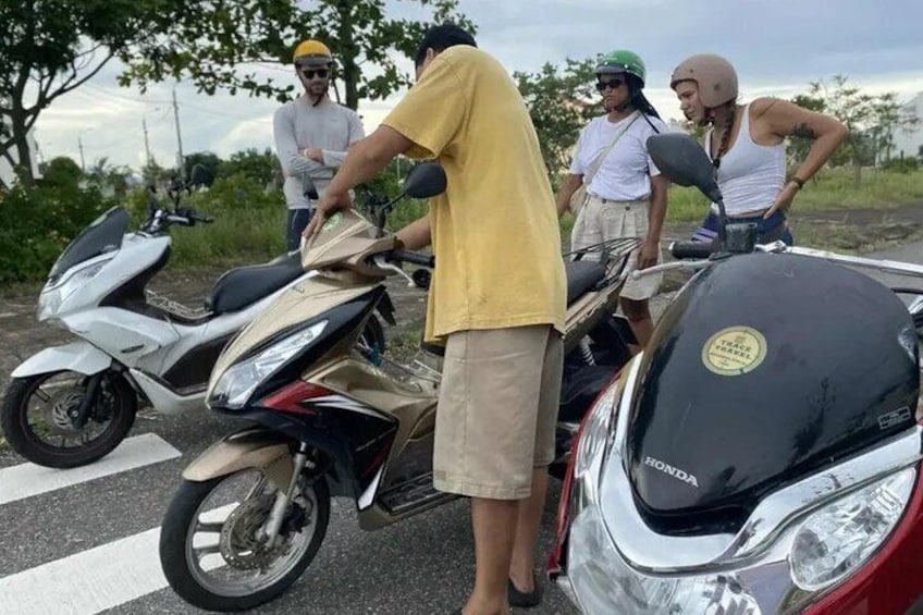 En gruppe mennesker står ved siden af en hvid og en rød motorcykel, med et træ og en vej i baggrunden. 3