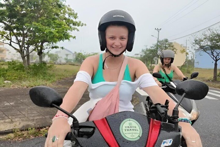 To personer på en motorcykel, hvoraf den ene bærer hjelm, samt en vej med træer og en klar himmel. 1