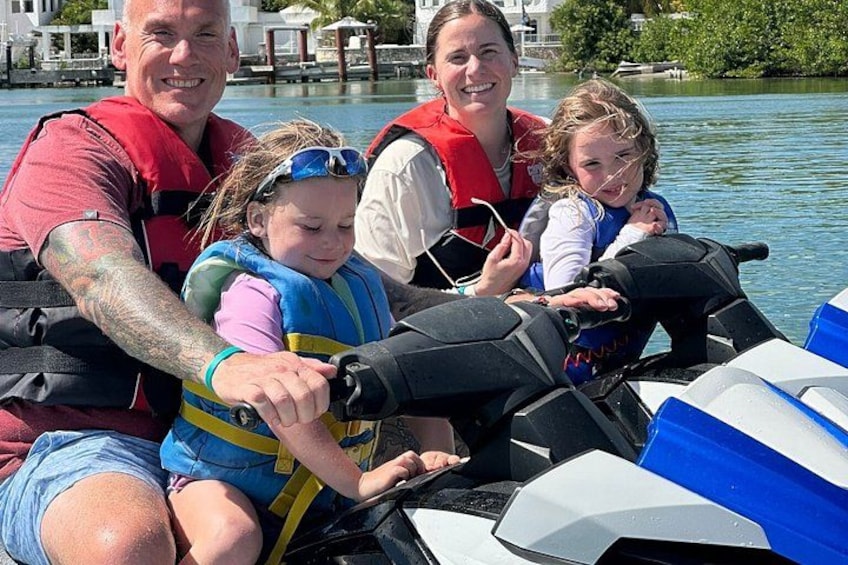 A group of people on a watercraft, wearing life jackets, with a body of water and buildings in the background. 3