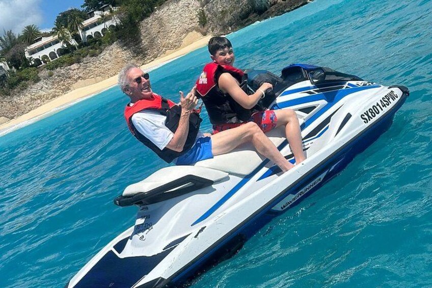 Two people on a jet ski with a beach and rocky shoreline in the background. 2